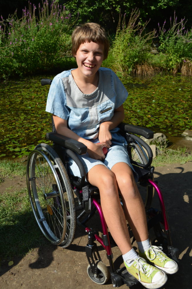 Dziewczynka na wózku inwalidzkim siedzi na tle oczka wodnego w którym znajdują się pływające rośliny wodne, w tle oczka rośliny i trawy ozdobne. 