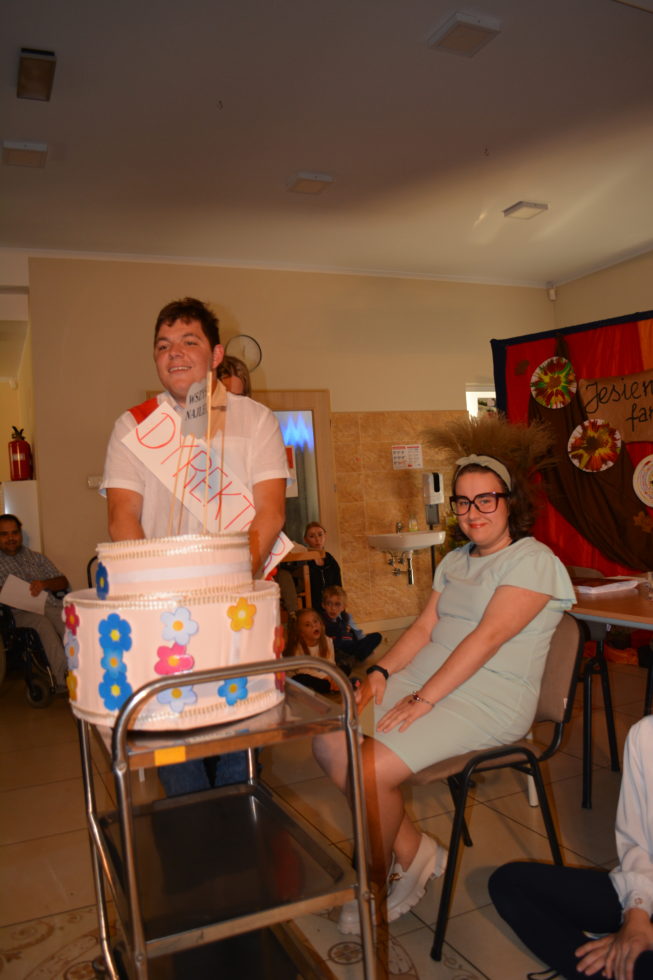 Na fotografii Paulina w roli nauczyciela siedzi na krześle, Daniel w roli dyrektora wiezie na wózku duży tort.