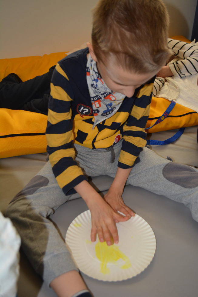 Na fotografii chłopiec maluje paluszkiem żółtą farbą słonko na papierowym talerzyku.