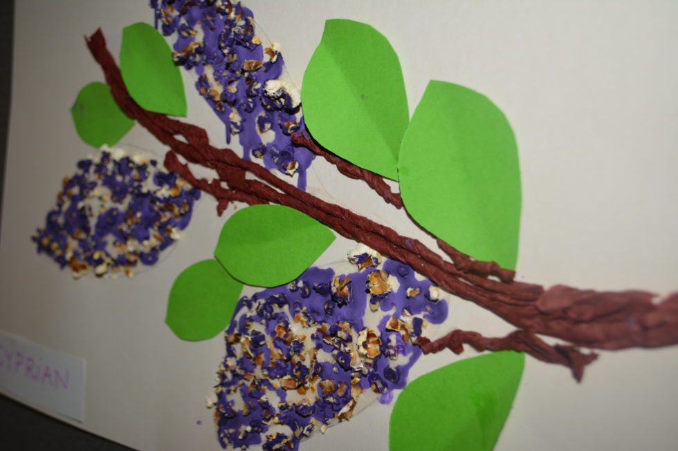 Na fotografii praca plastyczna przedstawiająca gałązkę bzu, wykonana z brązowej bibuły, fioletowej farby, popcornu i zielonych listków z papieru.