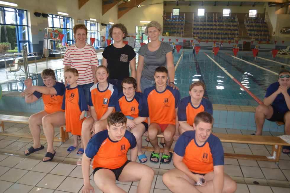 Na zdjęciu uczniowie siedzą na ławce na krytej pływalni wraz z opiekunami w trakcie zawodów pływackich.