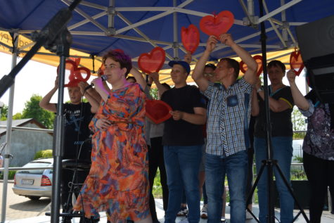 Na fotografii Paulina śpiewa piosenkę do mikrofonu na scenie, za nią koledzy i koleżanki trzymają czerwone serca.