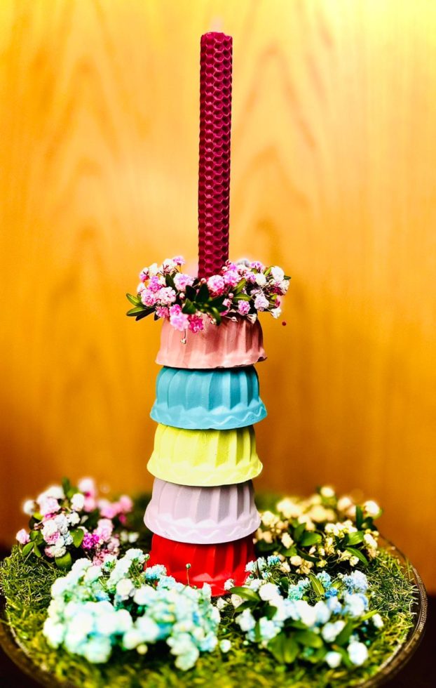 Na zdjęciu kolorowe babeczki wykonane z gipsu ustawione jedna na drugiej, na szczycie świeczka w kolorze fuksjowym z wosku pszczelego oraz wianuszek z różowej gipsówki.