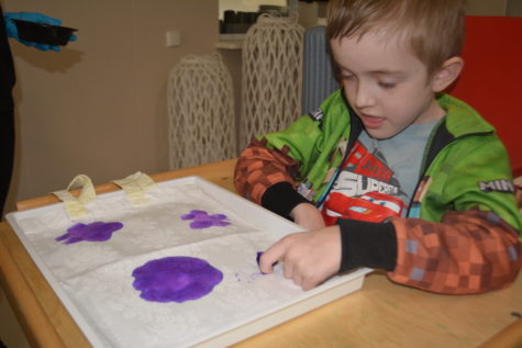 Na fotografii chłopiec siedzi w krzesełku i mokrą kolorową fioletową bibułą robi kropki na płóciennej torbie.