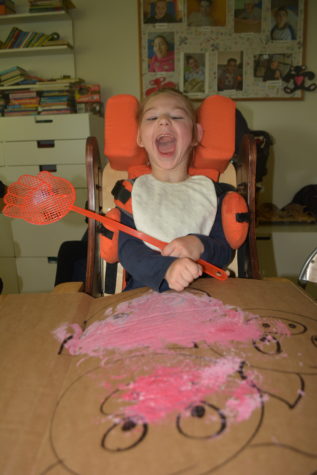 Na zdjęciu dziewczynka w krzesełku maluje farbami i packa na muchy obrazek przedstawiający trzy świnki.