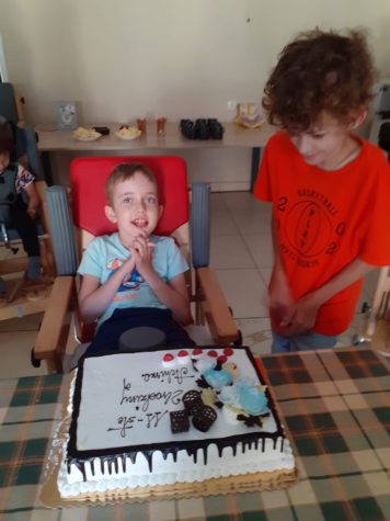 Na zdjęciu Achim siedzi w krzesełku, obok stoi kolega a przed nim tort urodzinowy.