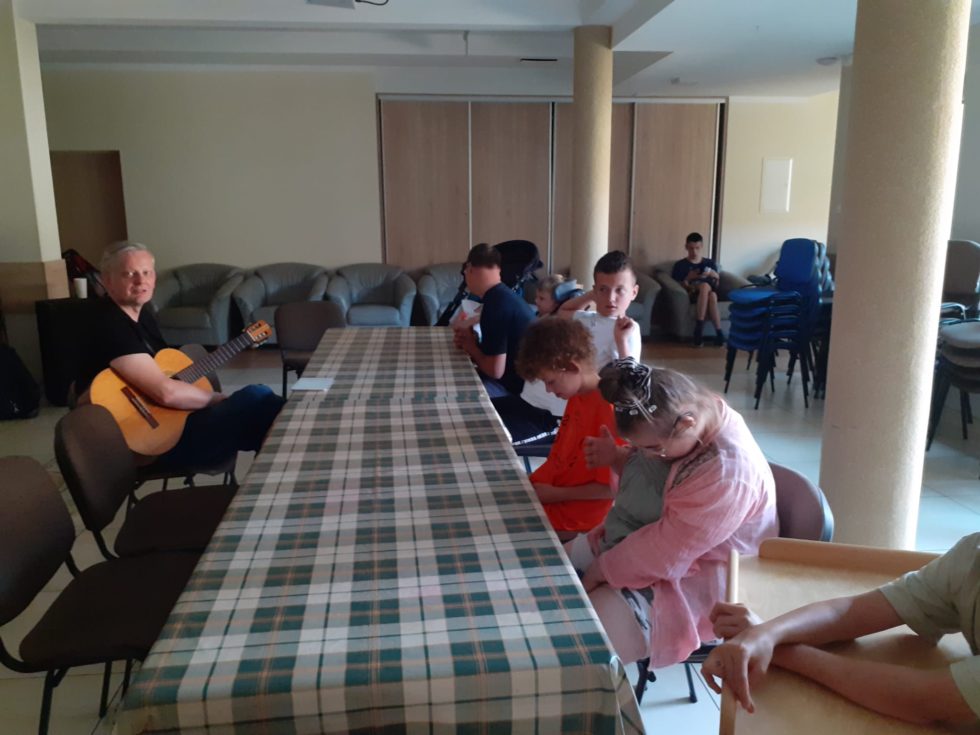 Na zdjęciu Pan z gitarą i dzieci siedzą przy stole.