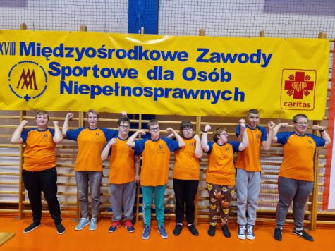 Na fotografii uczniowie w strojach sportowych, pomarańczowych koszulkach na tle baneru z napisem osiemnaste międzyośrodkowe zawody sportowe dla osób niepełnosprawnych.