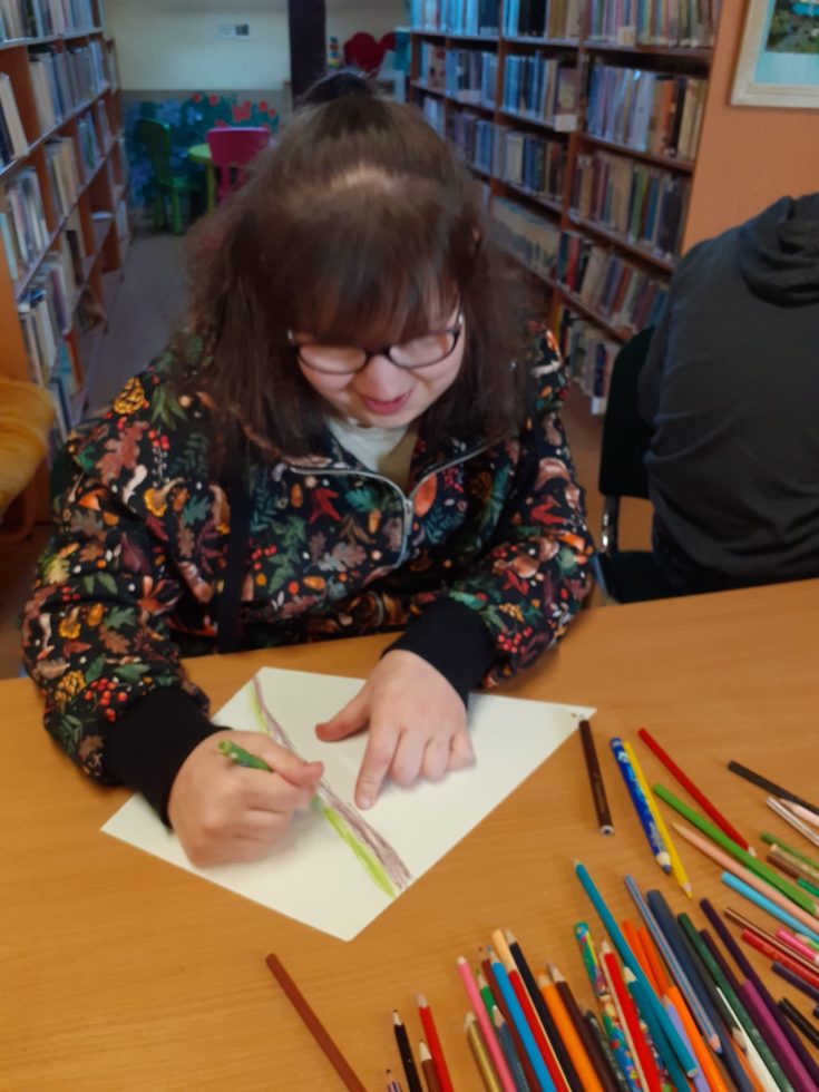 Na fotografii dziewczynka koloruje kolorowymi kredkami kartkę w kształcie latawca podczas zajęć bibliotecznych. W tle regały z książkami.