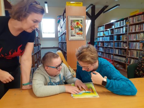 Na zdjęciu uczniowie oglądają obrazek obok stoi Pani bibliotekarka w tle regały z książkami.