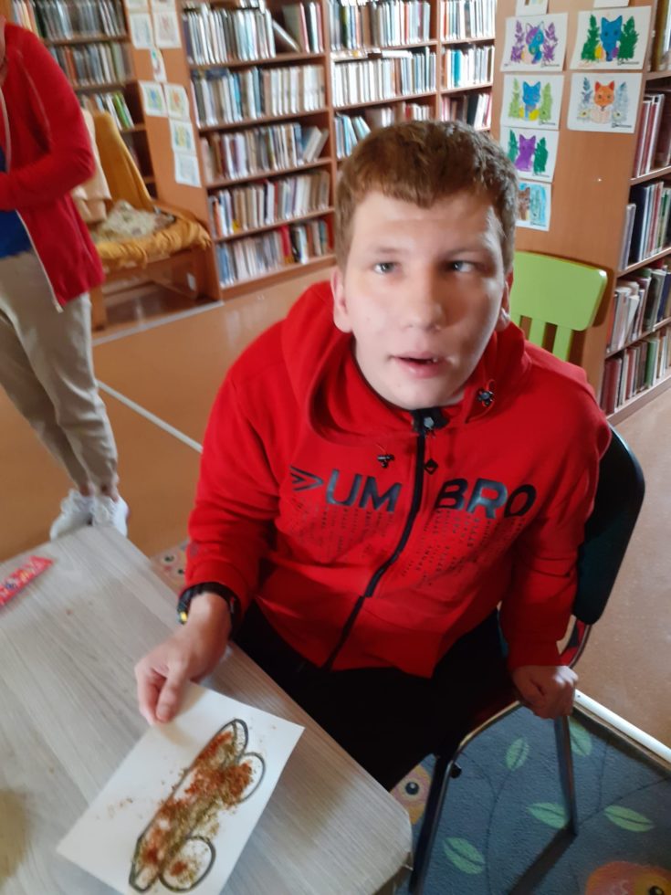 Na zdjęciu chłopiec siedzi przy stole w tle regały z książkami, trzyma obrazek który wykonał podczas zajęć bibliotecznych.