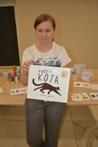 Na zdjęciu stoi Pani bibliotekarka trzyma w ręku książkę na okładce jest czarny kot.