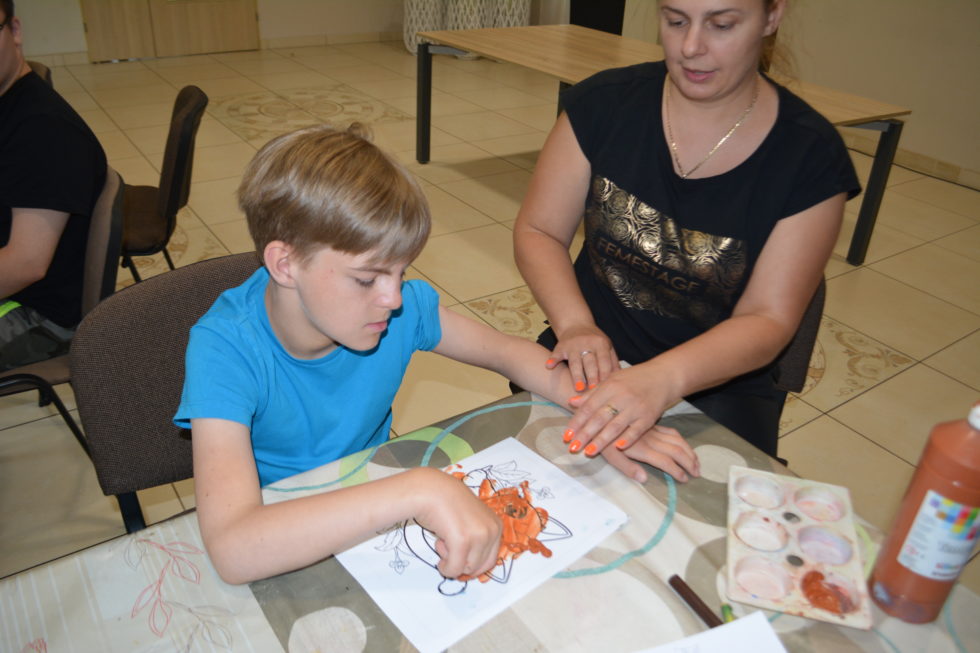 Na fotografii przy stole siedzi chłopiec i Pani. Chłopiec maluje obrazek ręką z użyciem farby, Pani mu pomaga.