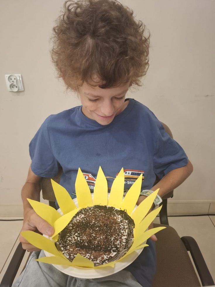 Na zdjęciu chłopiec siedzi na krześle , rękami trzyma duży słonecznik wykonany z papieru i brązowych ziarenek podczas zajęć plastycznych- bibliotecznych.