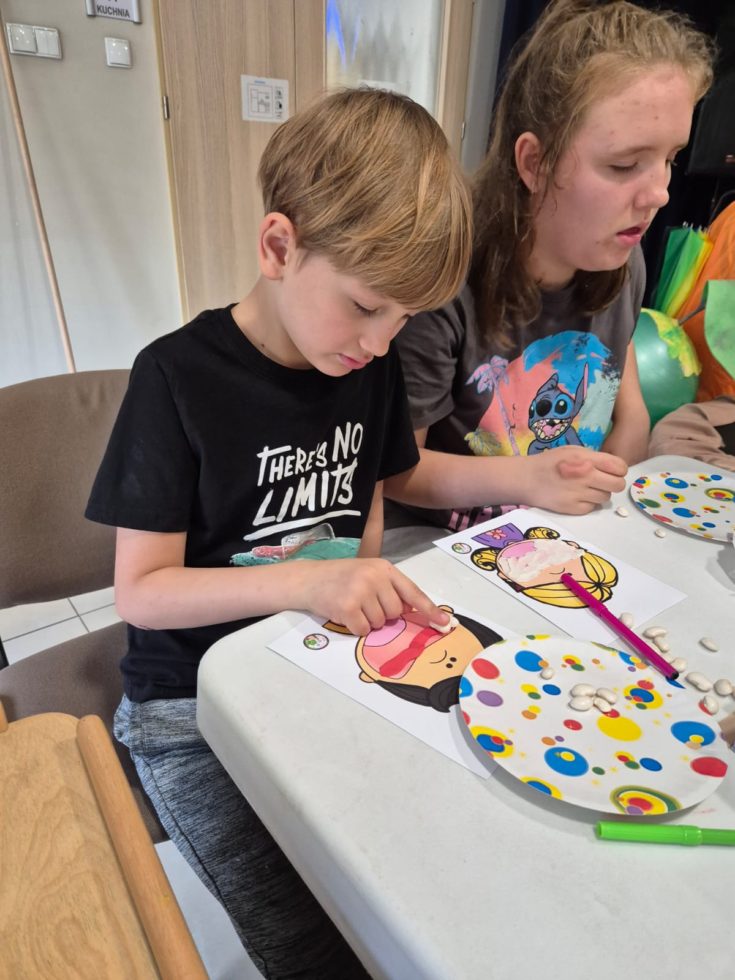 Na zdjęciu chłopiec i dziewczynka siedzą przy stole, chłopiec przykleja białe fasolki do plasteliny na obrazek który przedstawia otwarta buzię dziecka. Fasolki to zęby, plastelina to dziąsła.