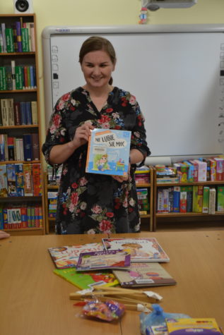 Na zdjęciu kobieta, bibliotekarka, stoi przy stole, trzyma książkę w rękach. Na stole leżą książki w tle regały z książkami podczas zajęć bibliotecznych.