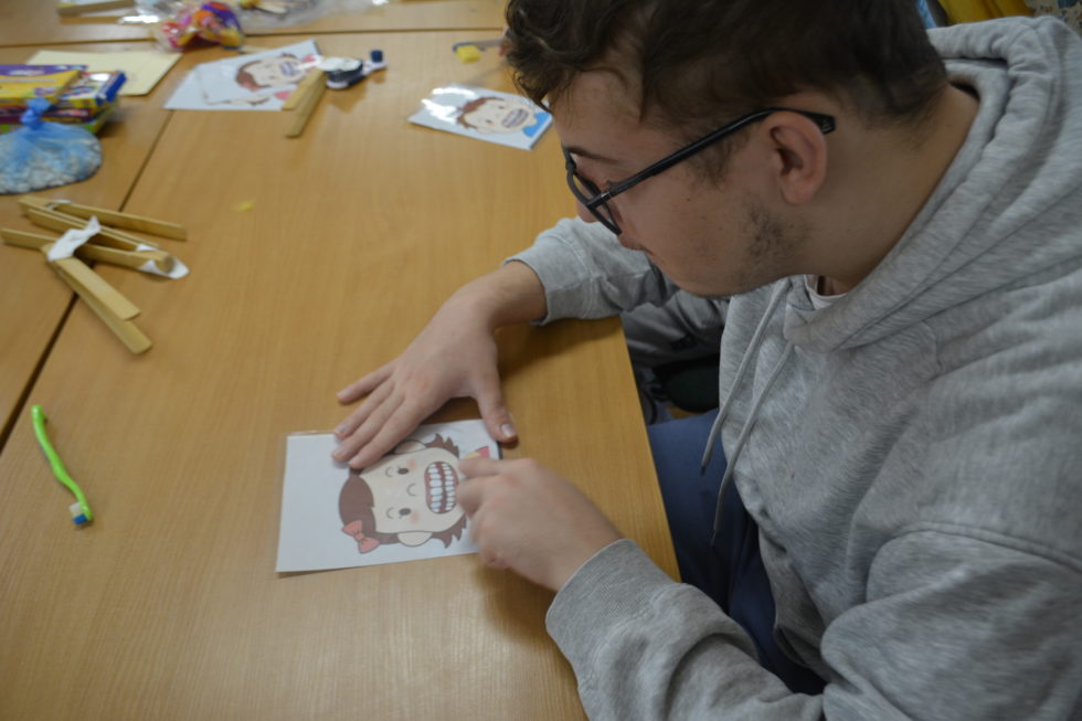 Na zdjęciu chłopak siedzi przy stole. Przed sobą ma obrazek dziewczynki z otwarta buzią. Chłopak ćwiczy szczotkowanie zębów.