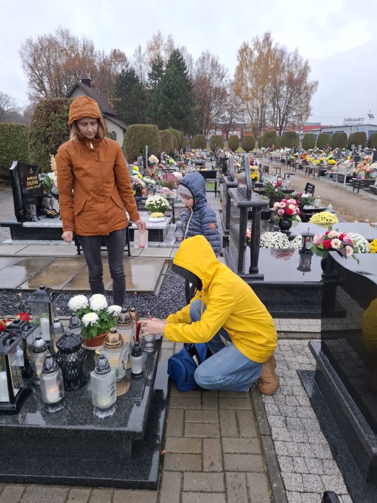 Na zdjęciu osoba w żółtej kurtce i założony kapturze stawia znicz na pomniku obok stoi chłopiec w kurtce z kapturem na głowie i opiekunka. W tle widać cmentarz z grobami.