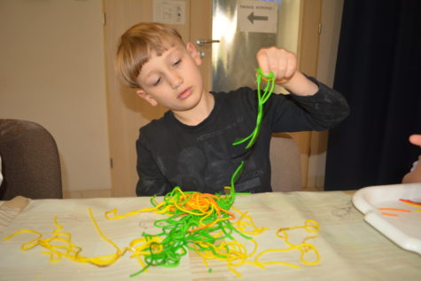 Na zdjęciu chłopiec siedzi przy stole przykrytym ceratą i bawi się zielonym ugotowanym makaron typu spagetii podczas warsztatów na Dzień Makaronu.