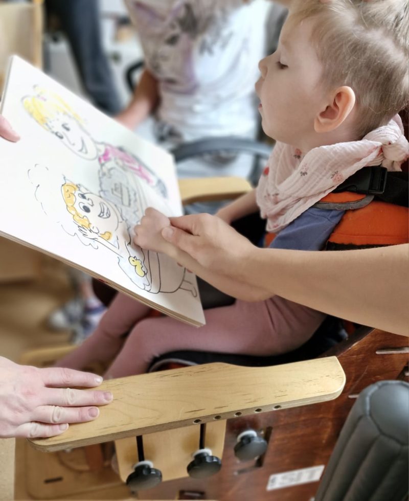 Mała dziewczynka siedzi w krzesełku rehabilitacyjnym i patrzy na ilustrację z książki. Opiekunka trzyma jej lewą rączkę i dotyka obrazka.