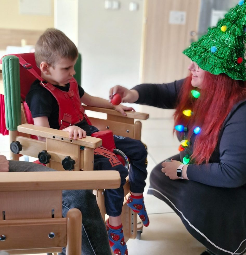 Na zdjęciu mały chłopiec siedzi w krzesełku terapeutycznym, przy nim kuca kobieta, na głowie ma zieloną czapkę w kształcie choinki z kolorowymi pomponami imitującymi bombki. Na szyi ma założone kolorowe świecące choinkowe lampki, prawej ręce trzyma czerwoną grzechotkę w kształcie jajka. Zdjęcie wykonane podczas warsztatów muzycznych.