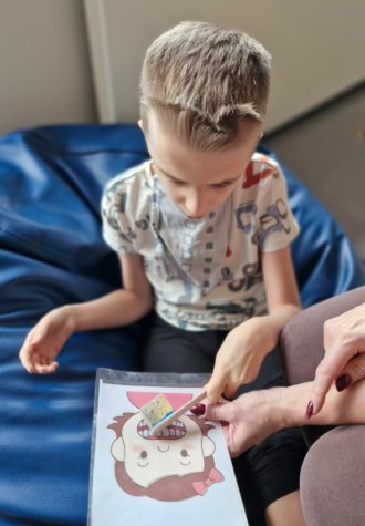 Na zdjęciu chłopiec siedzi na pufie przed sobą ma obrazek przedstawiający głowę dziecka z zębami. Chłopiec szczotkuje zęby na obrazku szczotką wykonana z patyczka i kawałka gąbki do mycia naczyń.