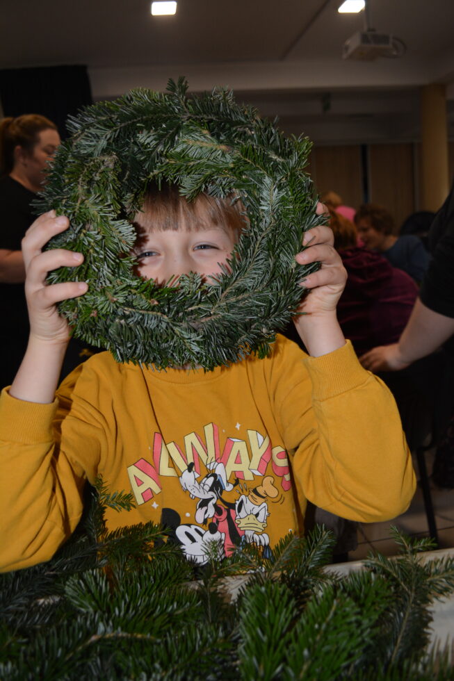 Na zdjęciu chłopiec siedzi przy stole, trzyma wianek z żywej zielonej jodły i zagląda przez otwór w wianku.
