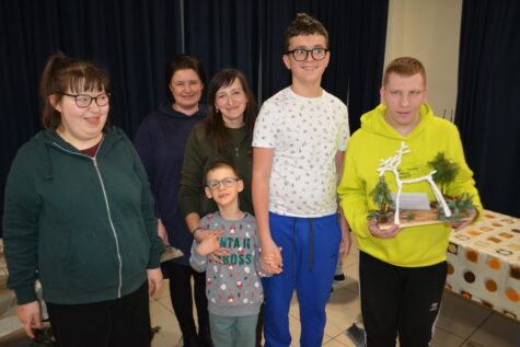 Na zdjęciu dwie kobiety, obok stoją trzej chłopcy i dziewczyna. Jeden z chłopców trzyma deskę z Reniferem z druta ozdobione zielonymi gałązkami.