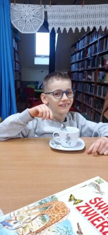 Na zdjęciu chłopiec w okularach siedzi przy stole i miesza łyżeczką w filiżance. W tle regały z książkami, u góry serwetki robione na szydełku.