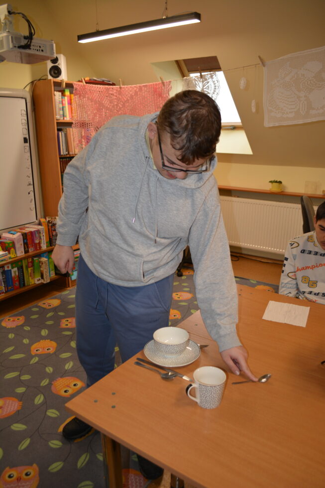 Na zdjęciu chłopak w okularach stoi przy stole, na stole stoi talerz na nim miska, obok kubek i ułożone sztućce. Podczas zajęć nakrywania stołu. W tle dywan i regały z książkami.