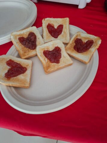 Na zdjęciu owalny papierowy talerz na którym leży pięć tostów, na tostach czerwone serduszka z dżemu truskawkowego. Pod talerzem czerwony obrus.