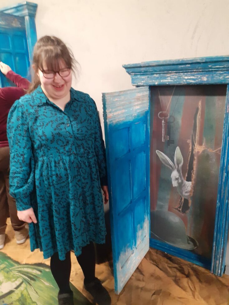 Na zdjęciu stoi dziewczyna w turkusowej sukience w czarne wzory, ma okulary. Obok niej na ścianie otwarte drzwiczki małej szafki w kolorze niebieskim przecierany białym. W środku szafki obraz przedstawiający zająca wychylającego się zza kurtyny, na dole namalowany melonik u góry klucz na sznurku. Zdjęcie zrobione w teatrze.