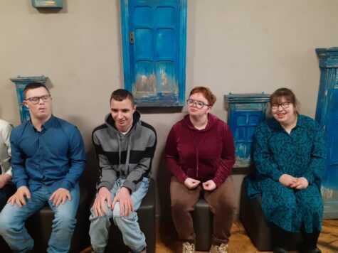 Na zdjęciu siedzą na szarych plastikowych krzesłach dwie dziewczyny obie w okularach i dwaj chłopcy, jeden w okularach. W tle na ścianie wiszą chabrowe skrzynki wyglądające jak okiennice. W oczekiwaniu na spektakl w teatrze.