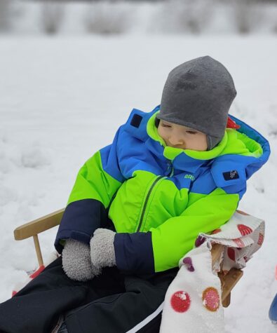 Na zdjęciu mały chłopiec w szarej czapce i kurtce w kolorze niebiesko zielonej siedzi na drewnianych sankach z oparciem. W tle biały śnieg.
