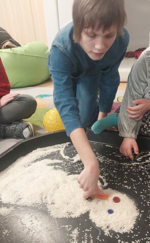 Na zdjęciu dziewczyna przykłada pomarańczowy nos bałwanowi zrobionemu z surowego ryżu. Bałwan jest na czarnej dużej tacy, oczy ma z guzików.