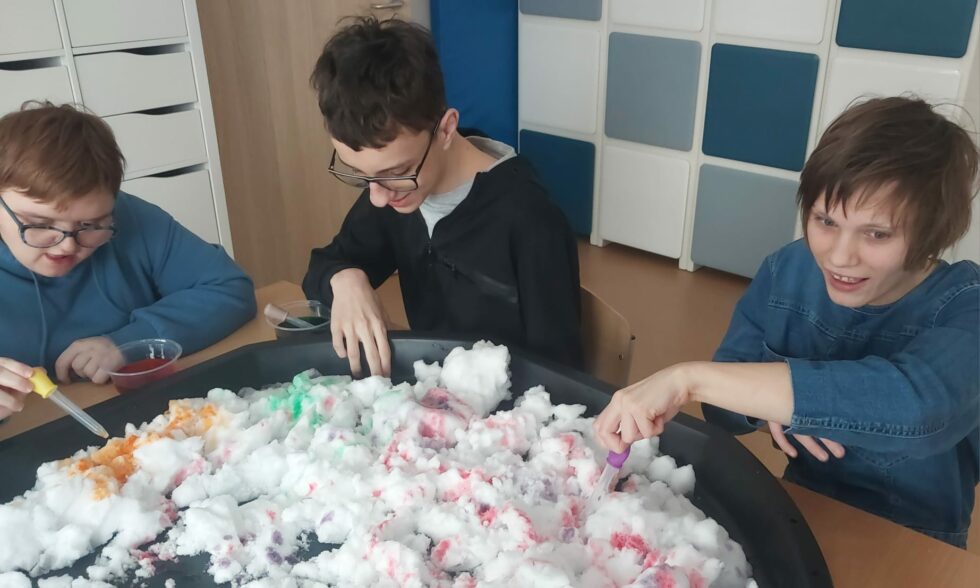 Na zdjęciu chłopak i dwie dziewczyny , jedna w okularach siedzą na krzesłach przy stole. Na stole leży duża czarna taca wypełniona śniegiem. Wychowankowie barwią śnieg kolorowymi płynami za pomocą pipety. Kolory płynów to: żółty, zielony i czerwony.