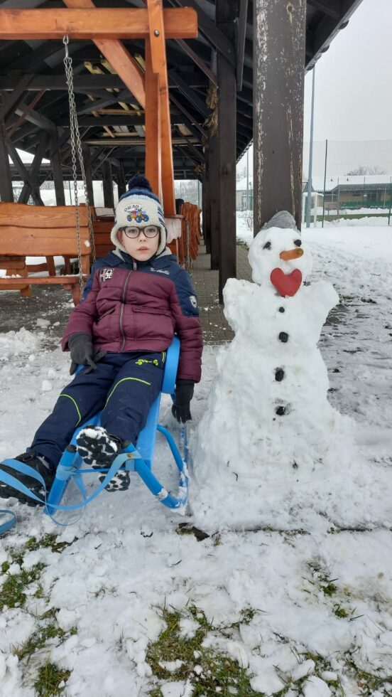 Na zdjęciu chłopiec w okularach w bordowo-granatowej kurtce i kolorowej czapce siedzi na niebieskich sankach. obok chłopca stoi mały bałwan ze śniegu. W tle wiata z drewnianymi ławkami.