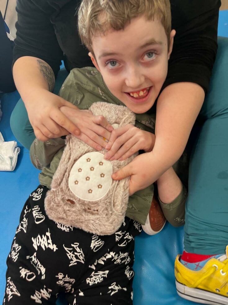 Na zdjęciu uśmiechnięty chłopiec leży na materacu i trzyma rękami termofor w kształcie sowy w kolorze beżowym. Widać również dłonie opiekunki , które pomagają mu trzymać termofor. Podczas zajęć bibliotecznych.