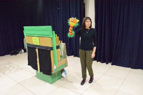 Na zdjęciu teatrzyk wykonany z pudełek tekturowych, z zasłoniętą kurtyną, obok stoi kobieta w okularach ma na prawej ręce założoną pacynkę przedstawiającą kolorowego smoka. W tle granatowe kotary.