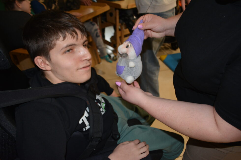 Na zdjęciu chłopak siedzi w wózku przypięty szelkami i z klinem dla osób niepełnosprawnych. Kobieta pokazuje mu marionetkę przedstawiającą skrzata w fioletowej czapce i szarych spodenkach z fioletową kieszonką.