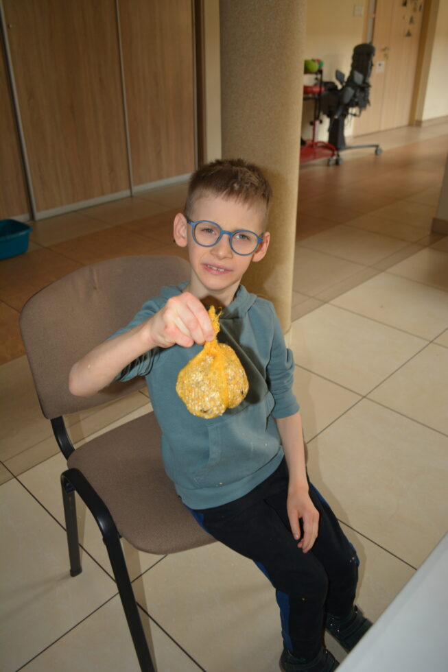 Na zdjęciu chłopiec w okularach siedzi na krześle. W prawej ręce trzyma pokarm dla ptaków w żółtej siatce.