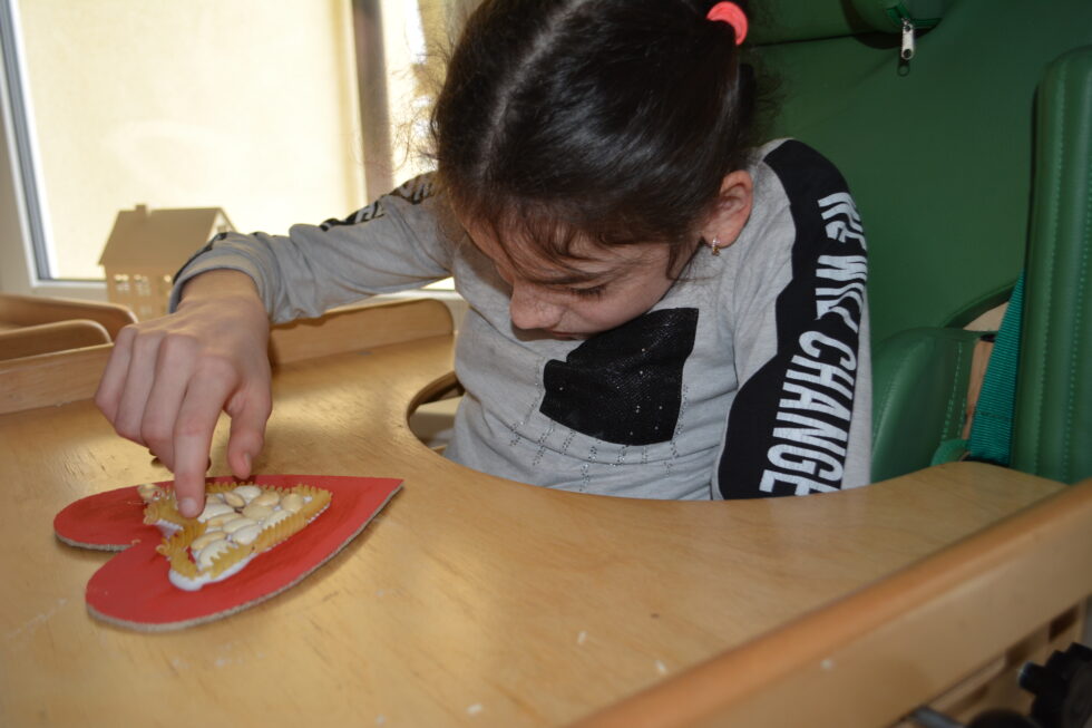 Na zdjęciu dziewczynka siedzi w krzesełku terapeutycznym, na blacie leży papierowe czerwone serce. Dziewczynka dekoruje serce makaronem świderkami i białą fasolą.