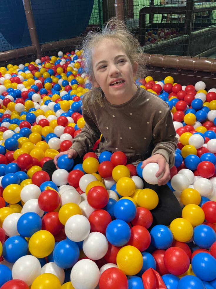 Na zdjęciu dziewczynka siedzi w kolorowych piłkach, w prawej ręce trzyma niebieską piłkę w lewej ręce białą.