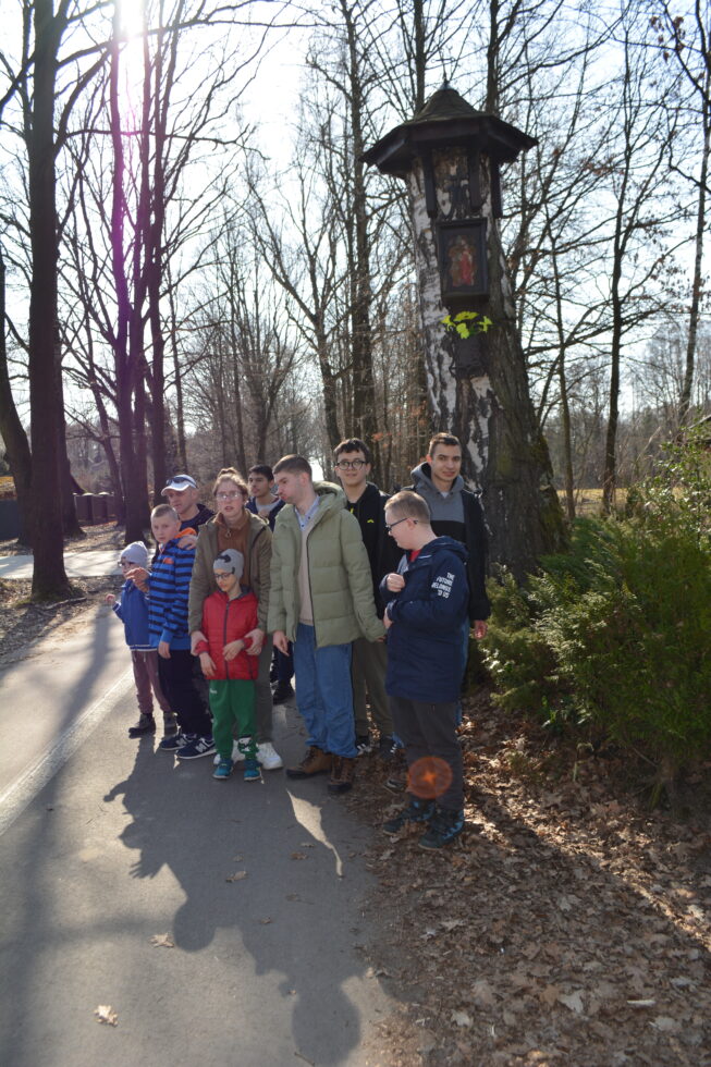 Chłopcy i dziewczyny wraz z opiekunem stoją przy drzewie na którym zawieszona jest kapliczka. Wewnątrz kapliczki stara figura przedstawiająca Jezusa. W tle aleja z drzewami.