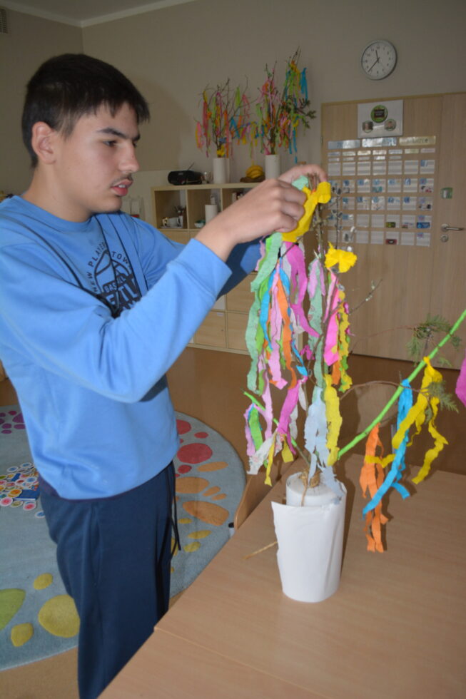 Na zdjęciu stoi chłopak przy stole. Na stole stoją związane gałązki wsadzone w stojak. Chłopak ozdabia gałązki kolorową bibułą.