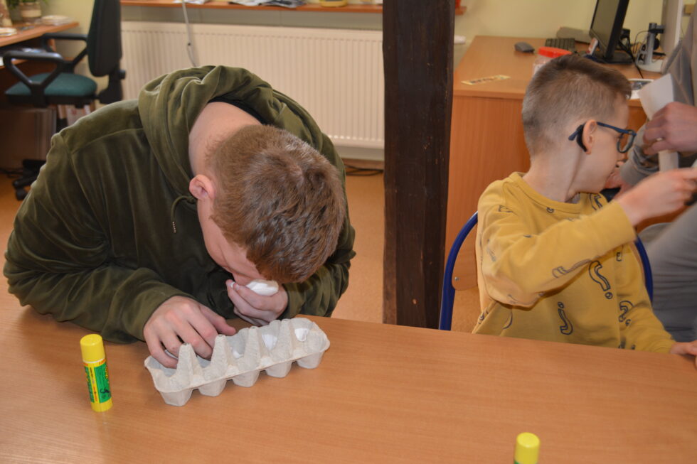 Na zdjęciu chłopak siedzi przy stole, do wytłaczanki na jajka wkłada po jednym płatku kosmetycznym do dziurki. Obok siedzi na krześle drugi mniejszy chłopiec w okularach i odwraca się do tyłu. 