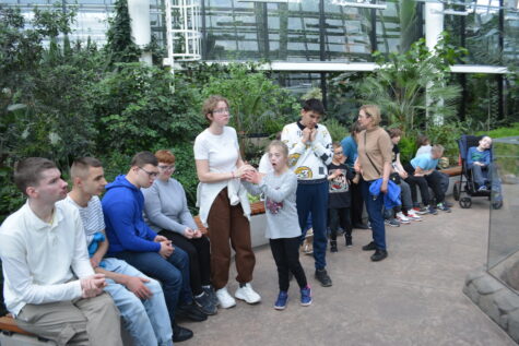 Na zdjęciu chłopcy i dziewczyny siedzą na długiej ławce, w tle rośliny tropikalne - palmiarnia.