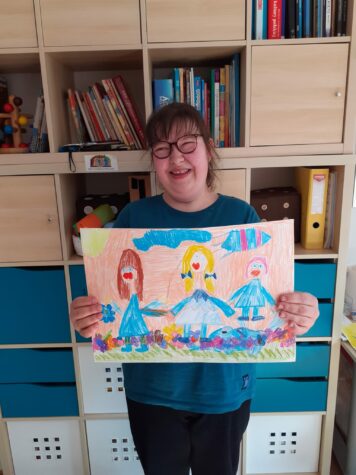 Na zdjęciu stoi dziewczyna w okularach trzyma pracę plastyczną przedstawiającą trzy postaci ubrane na niebiesko.