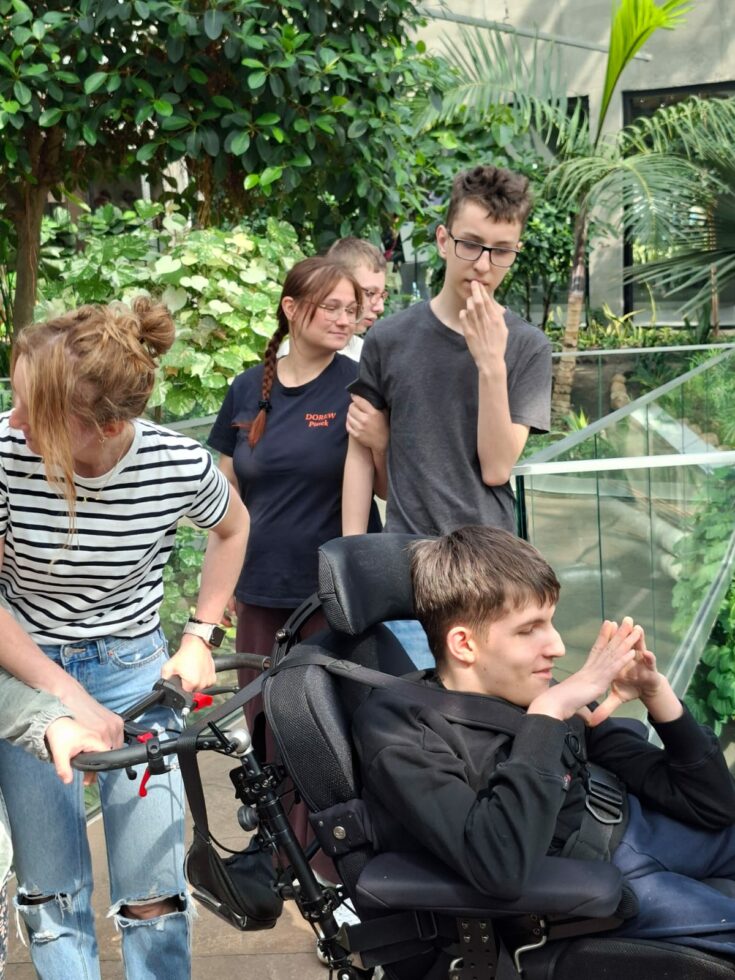 Na zdjęciu trzej chłopcy jeden w wózku terapeutycznym oraz dwie opiekunki. W tle palmiarnia z egzotycznymi roślinami.