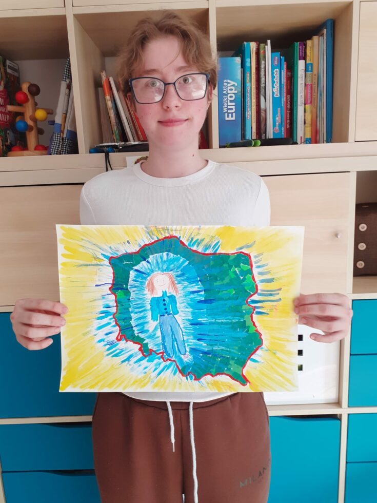 Na zdjęciu stoi dziewczyna w okularach trzyma obrazek na którym jest zarys Polski w środku postać ubrana na niebiesko.